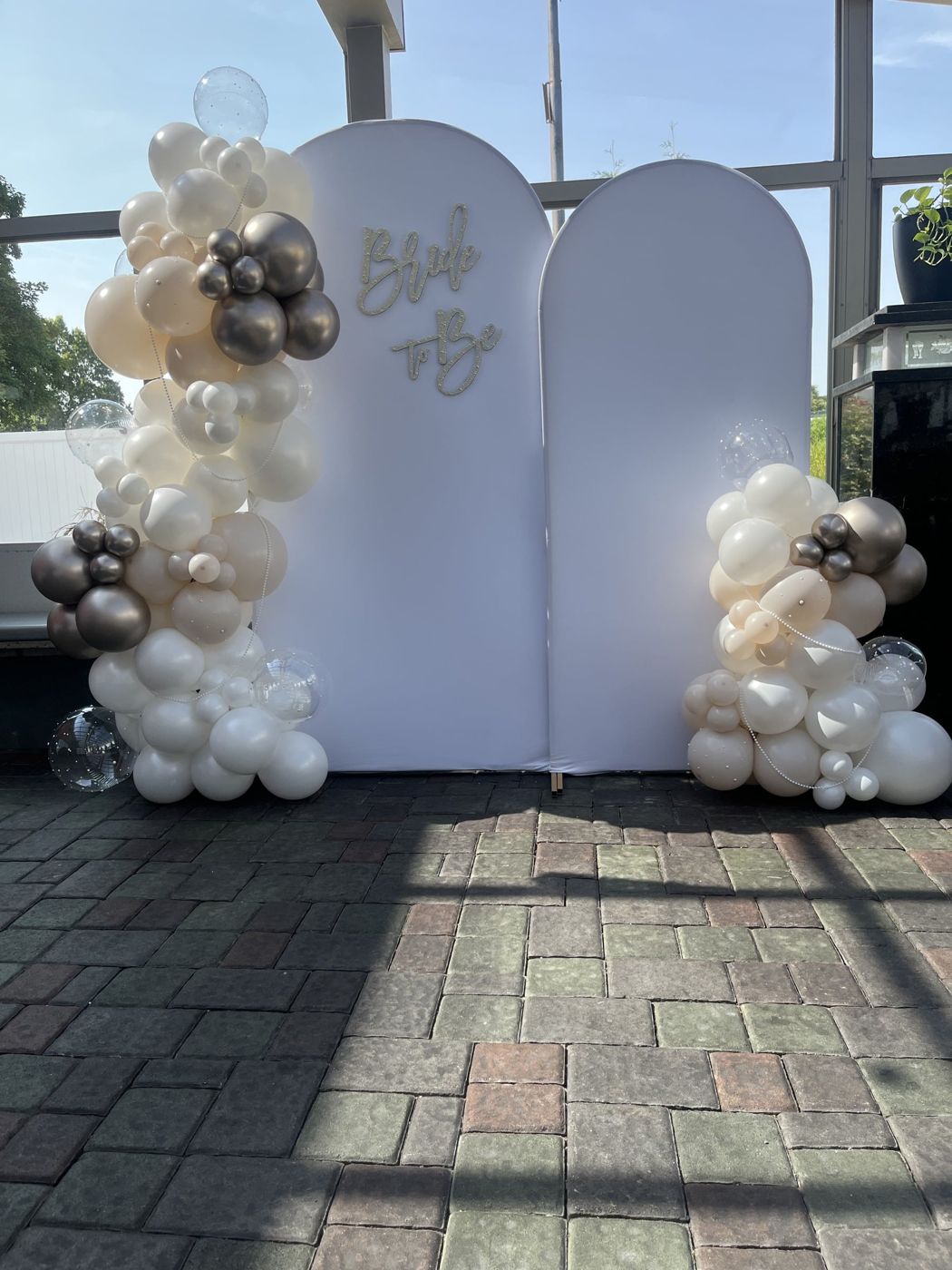 White wedding arch with neutral balloon garland on a brick patio.